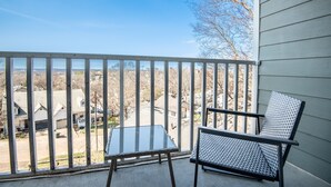 Un balcon avec une table en verre et deux chaises, donnant sur un quartier résidentiel avec des maisons et des arbres.