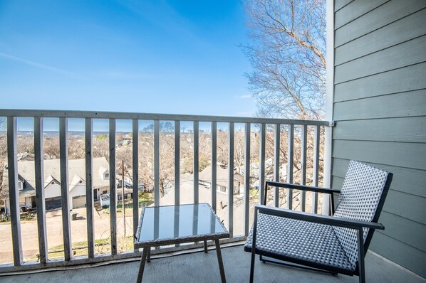 A balcony with a glass table and two chairs, overlooking a residential area with houses and trees.