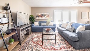 A living room with a blue sofa, a glass coffee table, a TV on a wooden stand, and a patterned rug. - Ridge Retreats Unit E- WiFi/4 Queen Beds/Groups (Birmingham)