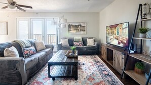 A living room with a dark gray sofa, a coffee table, and a patterned rug. There is a television on a wooden stand and a ceiling fan. - Ridge Retreats - Unit F: King Bed/Free Parking/TVs (Birmingham)