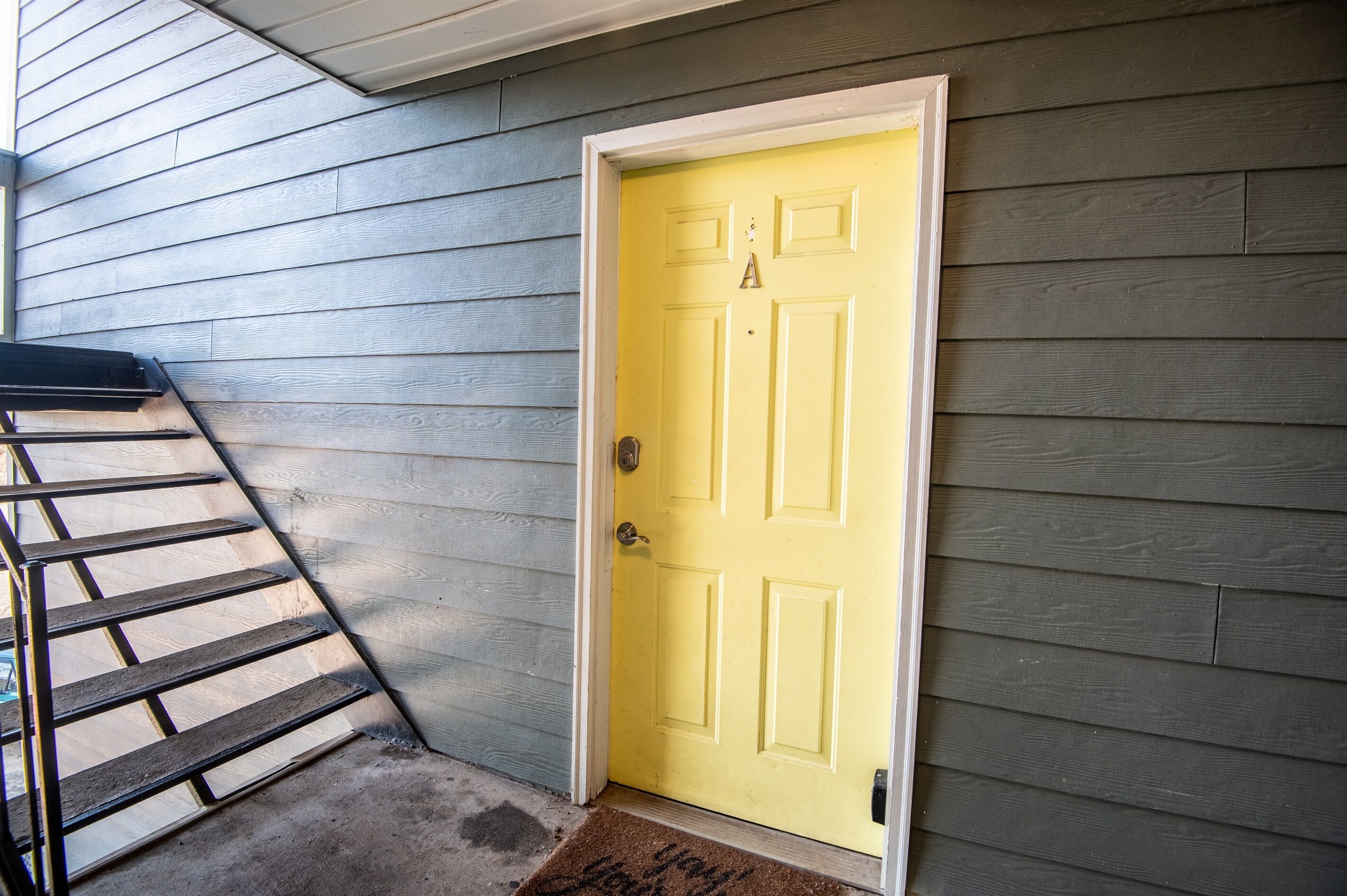 A yellow door with a white frame, a metal ladder leaning against a gray wooden wall, and a brown doormat with the word "Welcome".