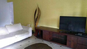 Una sala de estar con un sofá blanco, una mesa redonda de centro, un televisor sobre un mueble de madera y una planta decorativa.