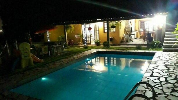 Una piscina con solárium empedrado, rodeada de mobiliario de exterior y un porche cubierto.