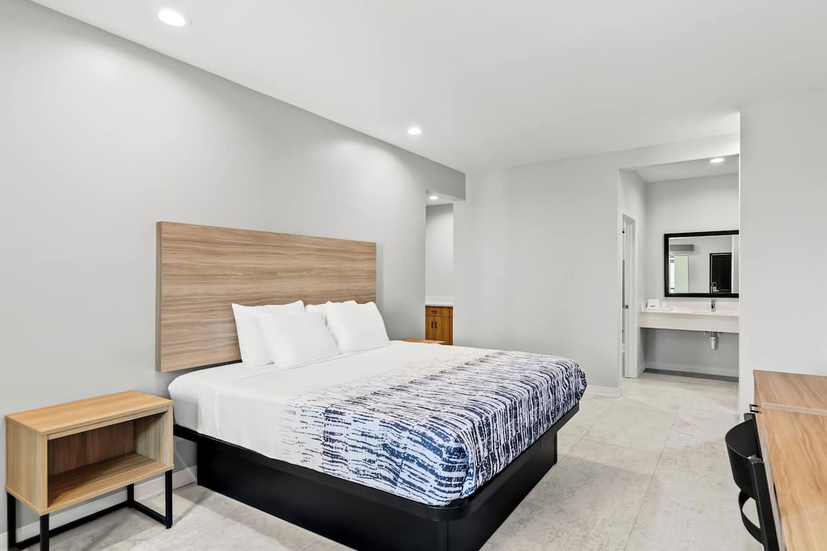 Une chambre d’hôtel moderne avec un grand lit, une tête de lit en bois, une table de chevet, un bureau et une salle de bain visible à travers une porte ouverte.