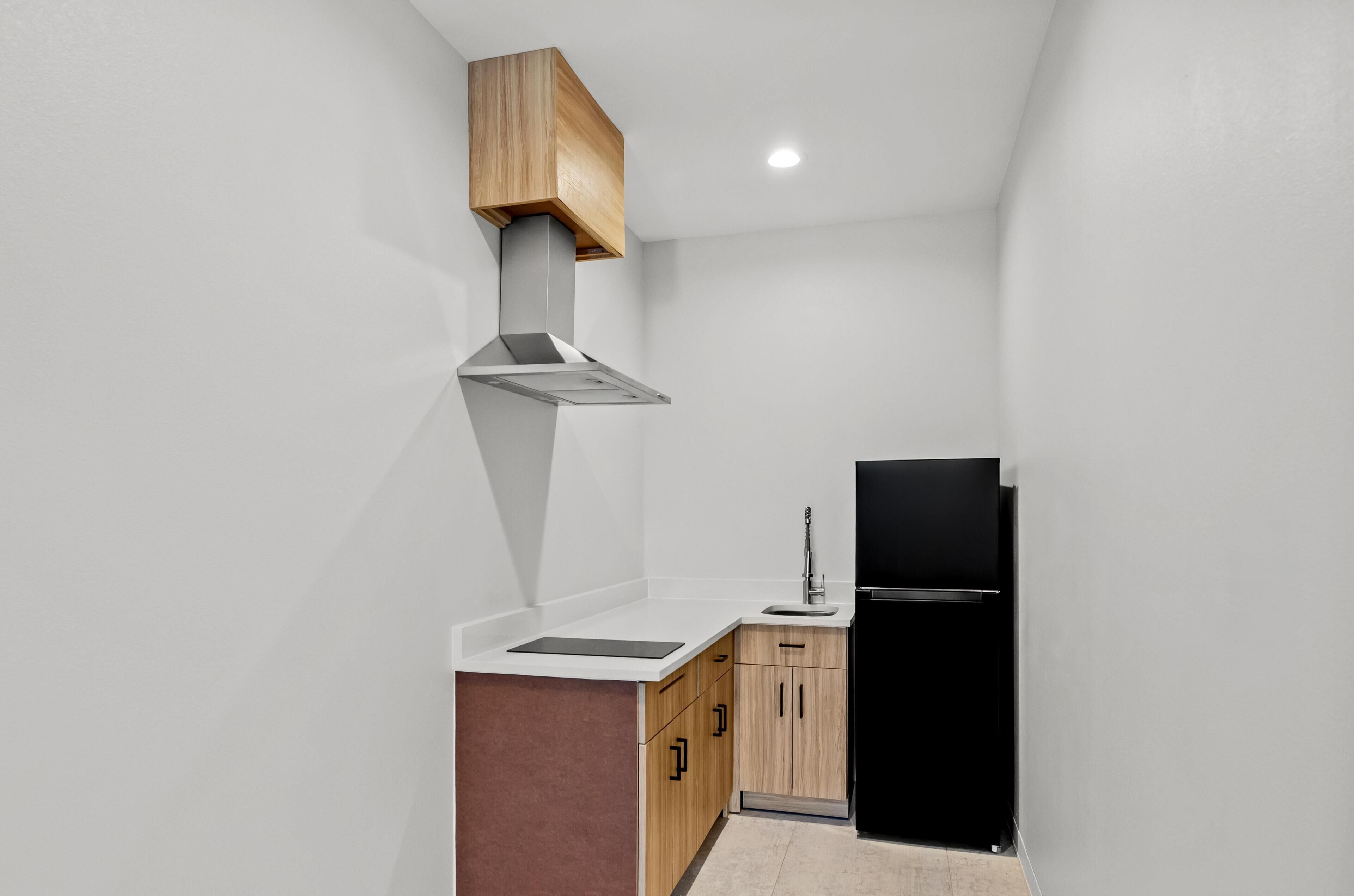 Eine kompakte Küche mit einem schwarzen Kühlschrank, Holzschränken, einer Edelstahl-Dunstabzugshaube und einer weißen Arbeitsplatte.