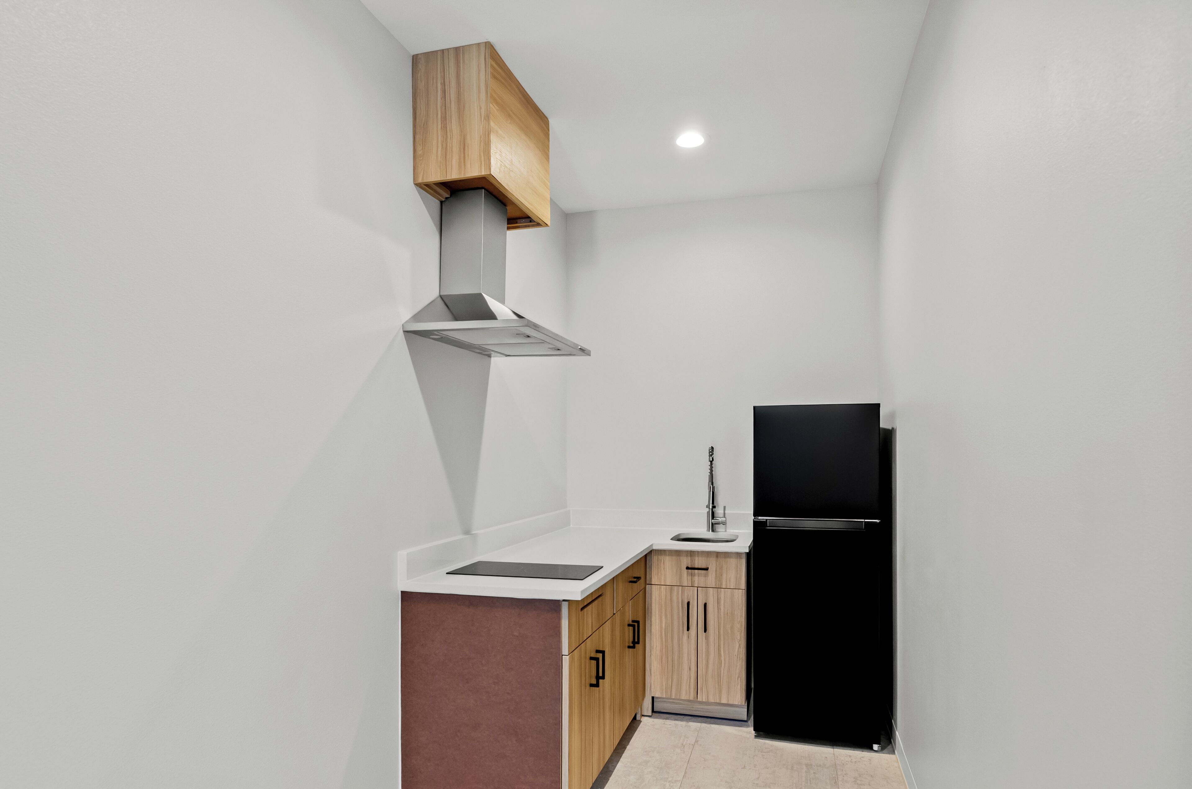 A compact kitchen with a black refrigerator, wooden cabinets, a stainless steel range hood, and a white countertop.