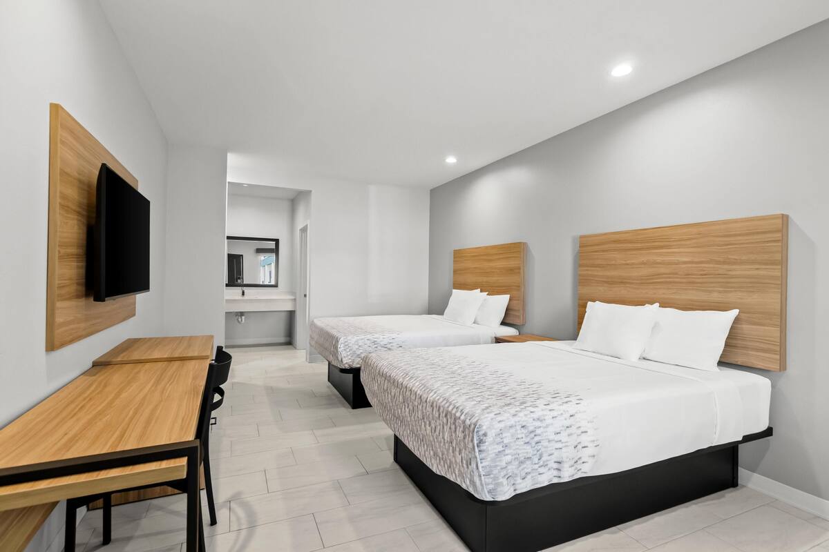 Une chambre d’hôtel moderne avec deux lits, un bureau en bois, un téléviseur à écran plat et une salle de bain visible à travers une porte ouverte.