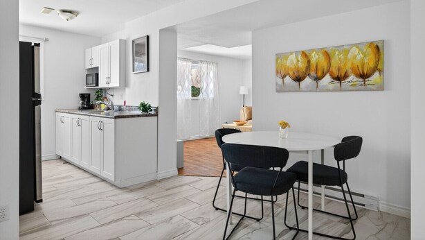 Eine moderne Küche mit weißen Schränken, einer Mikrowelle und einer Kaffeemaschine. Es gibt einen Essbereich mit einem weißen Tisch und schwarzen Stühlen, und ein Gemälde mit gelben Blumen an der Wand.