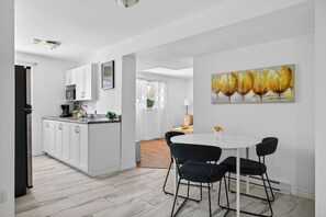 A modern kitchen with white cabinets, a microwave, and a coffee maker. There is a dining area with a white table and black chairs, and a painting of yellow flowers on the wall. - Charming Apartment by Aylmer Marina (Gatineau)