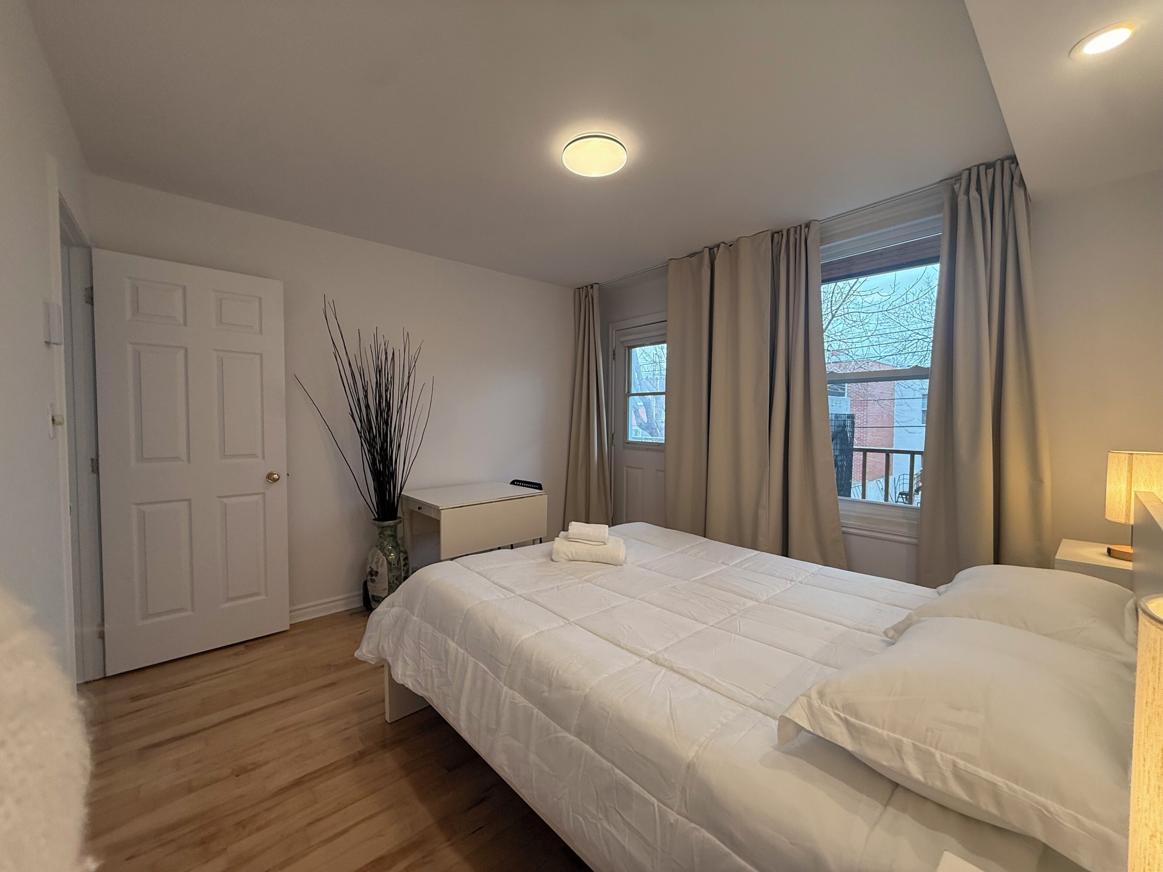 A bedroom with a bed, a bedside table, a lamp, a window with curtains, a door, and a vase with branches.