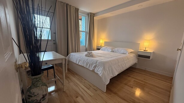 Ein Schlafzimmer mit einem Bett, Nachttischen, einem Schreibtisch, einem Stuhl, einem Fenster und einer Vase mit Zweigen.