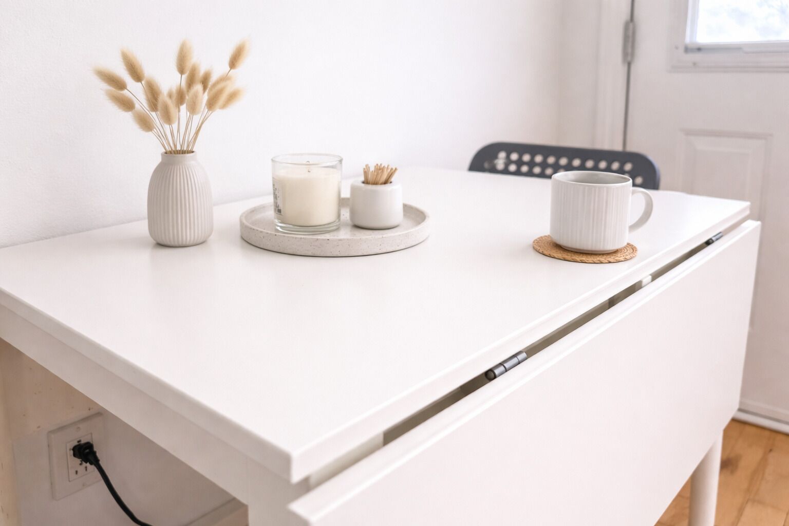 A white desk with a vase of pampas grass, a candle, and a mug.