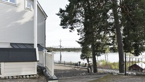 A house with a porch overlooking a lake, a gravel driveway, and a fenced area with a small cabin in the background.