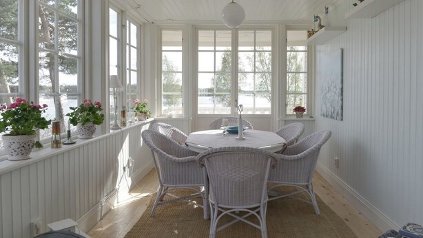 A bright, white-walled room with a round table and wicker chairs, large windows, and potted plants.