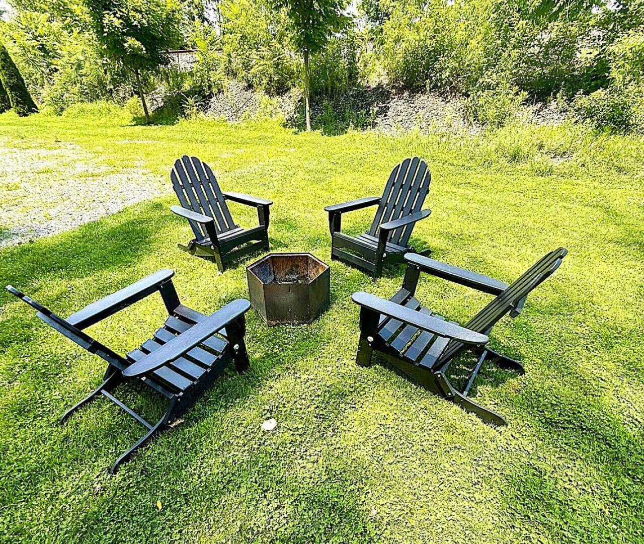 Four Adirondack chairs arranged in a circle around a fire pit on a grassy lawn with trees in the background.