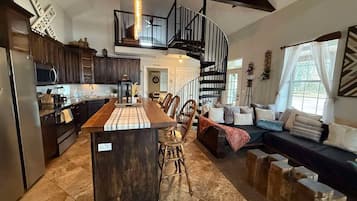 A spacious kitchen with a central island, a dining area with chairs, a staircase, and a living area with a sofa and cushions.