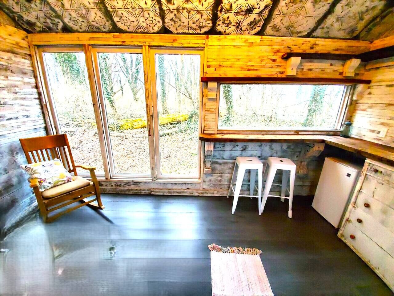 A cabin interior with a wooden chair, a stool, a small refrigerator, and large windows offering a view of the outdoors.