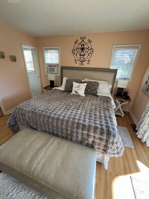 A bedroom with a large bed, a bench, two bedside tables, a wall decoration, and a window with blinds.