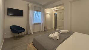 A modern hotel room with a flat-screen TV, a single blue chair, a bed with rolled towels, and a window with curtains. - Levante Apartment - Style and Elegance 300 meters from the sea (Santa Margherita di Pula)