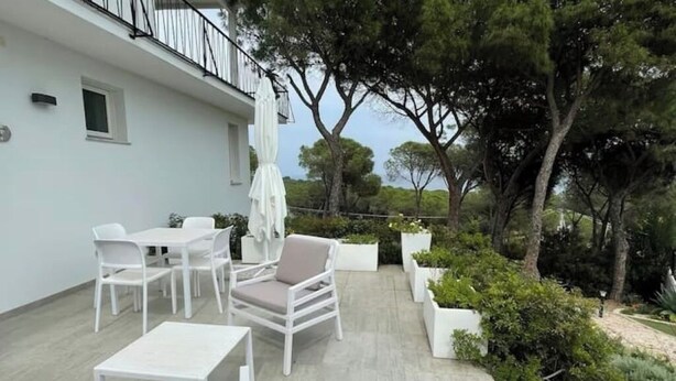 A patio with white furniture, a table, and chairs, surrounded by greenery and trees. - Levante Apartment - Style and Elegance 300 meters from the sea (Santa Margherita di Pula)
