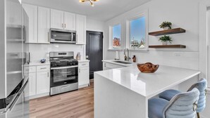 A modern kitchen with white cabinetry, stainless steel appliances, a central island, and a view of the outside through large windows. - Comfortable SFH for Families & Groups • Easy Walk to Local Shops and Eateries (Downers Grove)