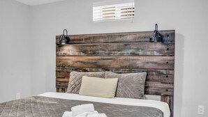 A bedroom with a wooden headboard, two wall-mounted lamps, a bed with a quilted bedspread, two pillows, and a pair of towels on the bed. - Spacious 6BR Full Home Near Niagara Falls & Wine (St. Catharines)