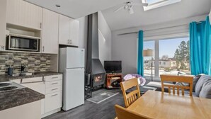 A modern kitchen with a built-in microwave, a refrigerator, and a wood-burning stove. There is a dining area with a wooden table and chairs, and a large window with curtains. - Condo MSA - Ski Beaupré (Beauprã©)