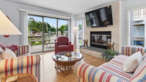 A living room with a fireplace, a TV mounted on the wall, and a large window overlooking a body of water. - Water's Edge Retreat OCMD (Ocean Pines)