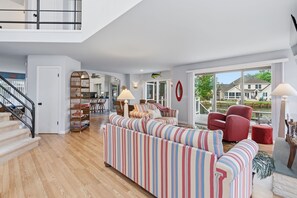 A spacious living room with a striped sofa, a red chair, and a staircase leading to a balcony. - Water's Edge Retreat OCMD (Ocean Pines)