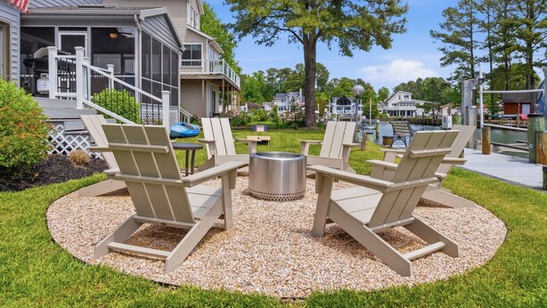 A patio with Adirondack chairs, a fire pit, and a view of a residential area with houses and a dock. - Water's Edge Retreat OCMD (Ocean Pines)