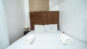 Een net opgemaakt bed met witte lakens en twee opgerolde handdoeken, een houten hoofdbord en een nachtkastje met een lamp.