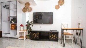 Een moderne woonkamer met een flatscreen televisie aan de muur, een zwarte entertainmentkast met gouden accenten, een witte boekenkast en een eethoek met een houten tafel en stoelen.