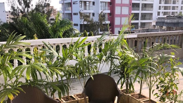 Een terras op het dak met twee rieten stoelen en een klein krukje, omgeven door potplanten en uitzicht op een stadsgezicht met hoge gebouwen.