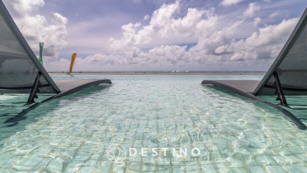 An infinity pool with clear water, two lounge chairs, and a view of the ocean under a partly cloudy sky. - APTO Térreo | 4 Quartos | Até 12 Pessoas | Barra Bali | Destino BSM 002 (Barra de São Miguel)
