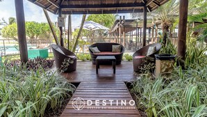 A covered outdoor seating area with wicker furniture and a pool in the background. - APTO Térreo | 4 Quartos | Até 12 Pessoas | Barra Bali | Destino BSM 002 (Barra de São Miguel)