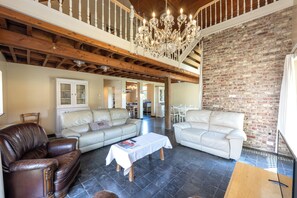 A spacious living room with a high ceiling, exposed wooden beams, a brick wall, and a chandelier. - Villa Krat (Holsbeek)