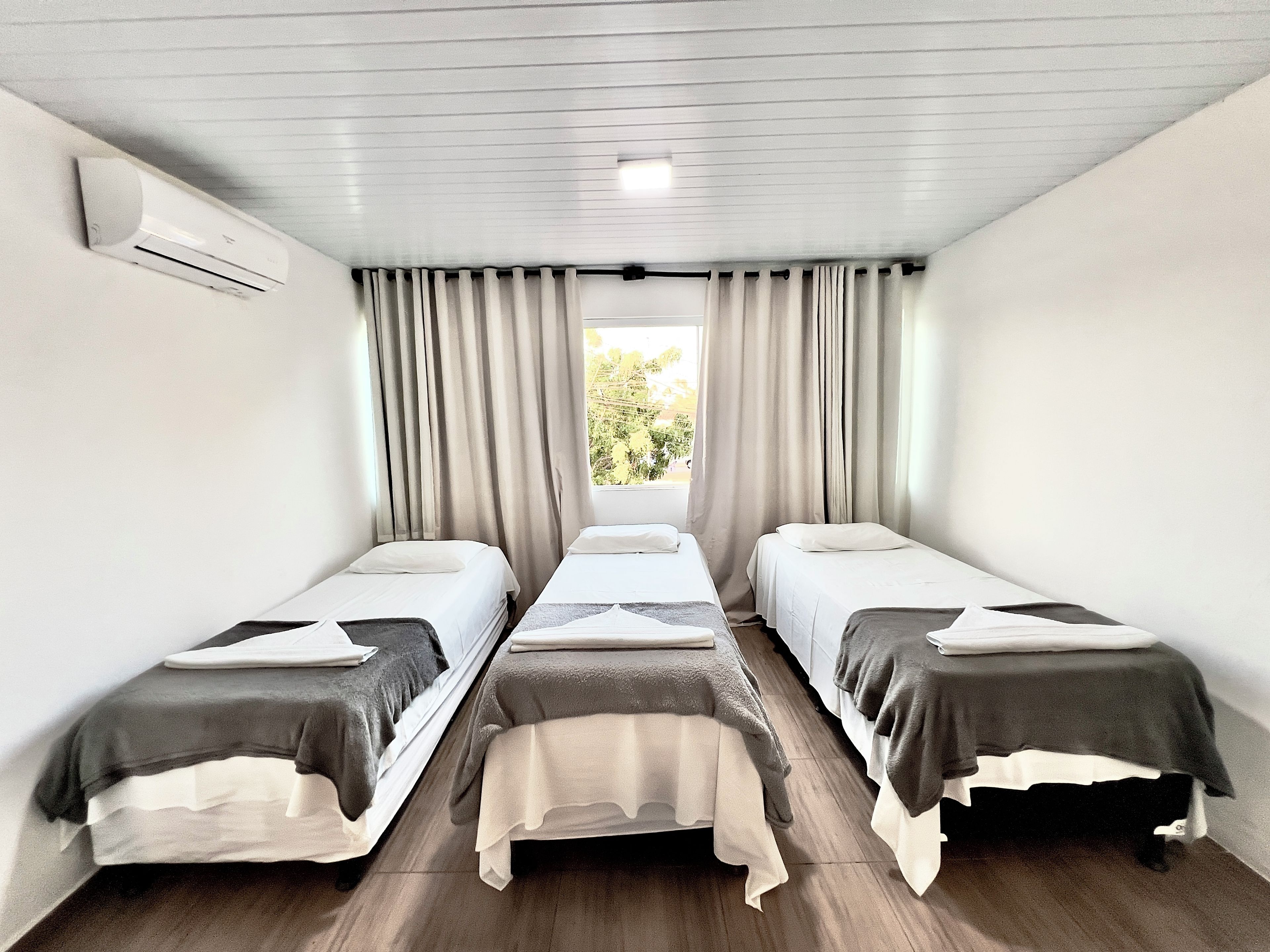 Habitación con tres camas individuales, una ventana con cortinas, un equipo de aire acondicionado y vistas a la vegetación.