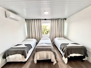 Habitación con tres camas individuales, una ventana con cortinas, un equipo de aire acondicionado y vistas a la vegetación.