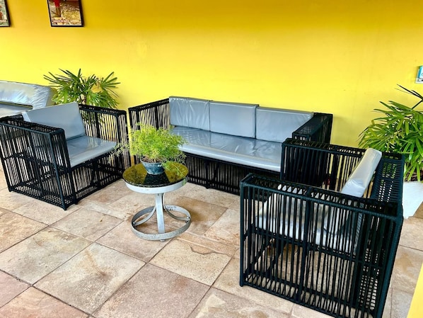 Un patio amueblado con un sofá modular, dos sillas de mimbre negro, una mesita redonda con una planta y plantas en macetas.