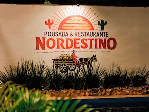 Un letrero con el texto 'POUSADA & RESTAURANTE NORDESTINO' y un gráfico de una persona en un carruaje tirado por caballos.