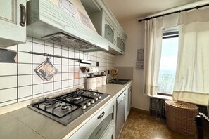 A kitchen with a gas stove, white tiled backsplash, and a window with curtains. - Mistral Apartment - Modern luxury and relaxation at a stone's throw from the sea (Santa Margherita di Pula)