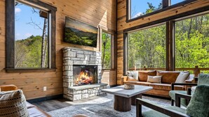 L’intérieur chaleureux d’un chalet avec une cheminée en pierre, des murs en bois, de grandes fenêtres et des sièges confortables.