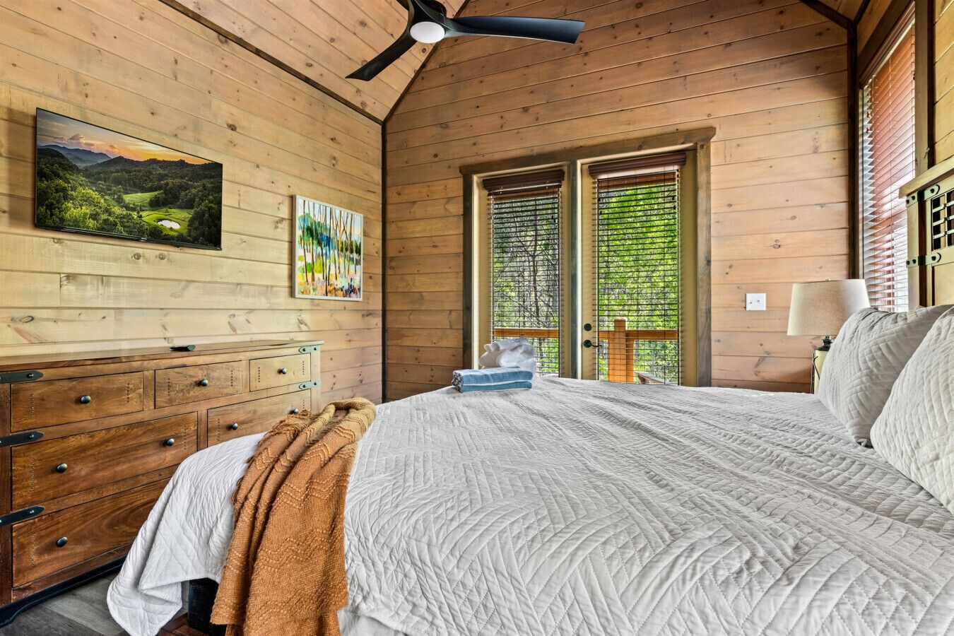 A wooden cabin bedroom with a bed, a dresser, a ceiling fan, a painting, and a window with blinds.
