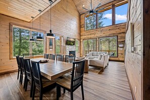 A wooden cabin interior with a dining area, a fireplace, and a sofa by a large window. - New! 5BR Pool Cabin w/Hot Tub*Theater*Game Room (Sevierville)