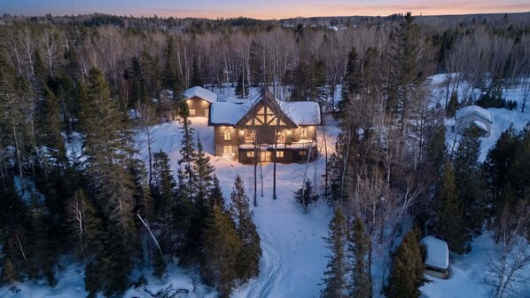 Une grande cabane en rondins à plusieurs étages, dotée d’une architecture symétrique, entourée d’une forêt dense et d’un sol recouvert de neige.