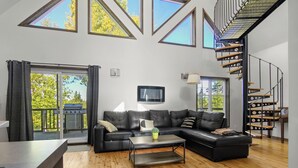 En moderne stue med en modulsofa, et sofabord og et fjernsyn monteret på væggen. Store vinduer og en trappe med et unikt design.