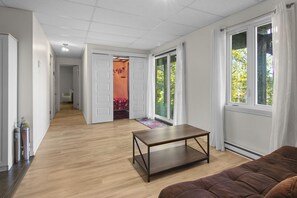 Un vaste espace de vie doté d’un plancher en bois, d’un canapé marron, d’une petite table et d’une porte-fenêtre coulissante donnant sur un balcon avec vue sur les arbres.