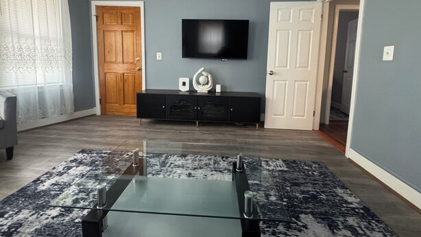 Un salon avec une table basse en verre, un téléviseur à écran plat fixé au mur, un meuble en bois foncé et un tapis à motifs.