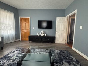 Un salon avec une table basse en verre, un téléviseur à écran plat fixé au mur, un meuble en bois foncé et un tapis à motifs.