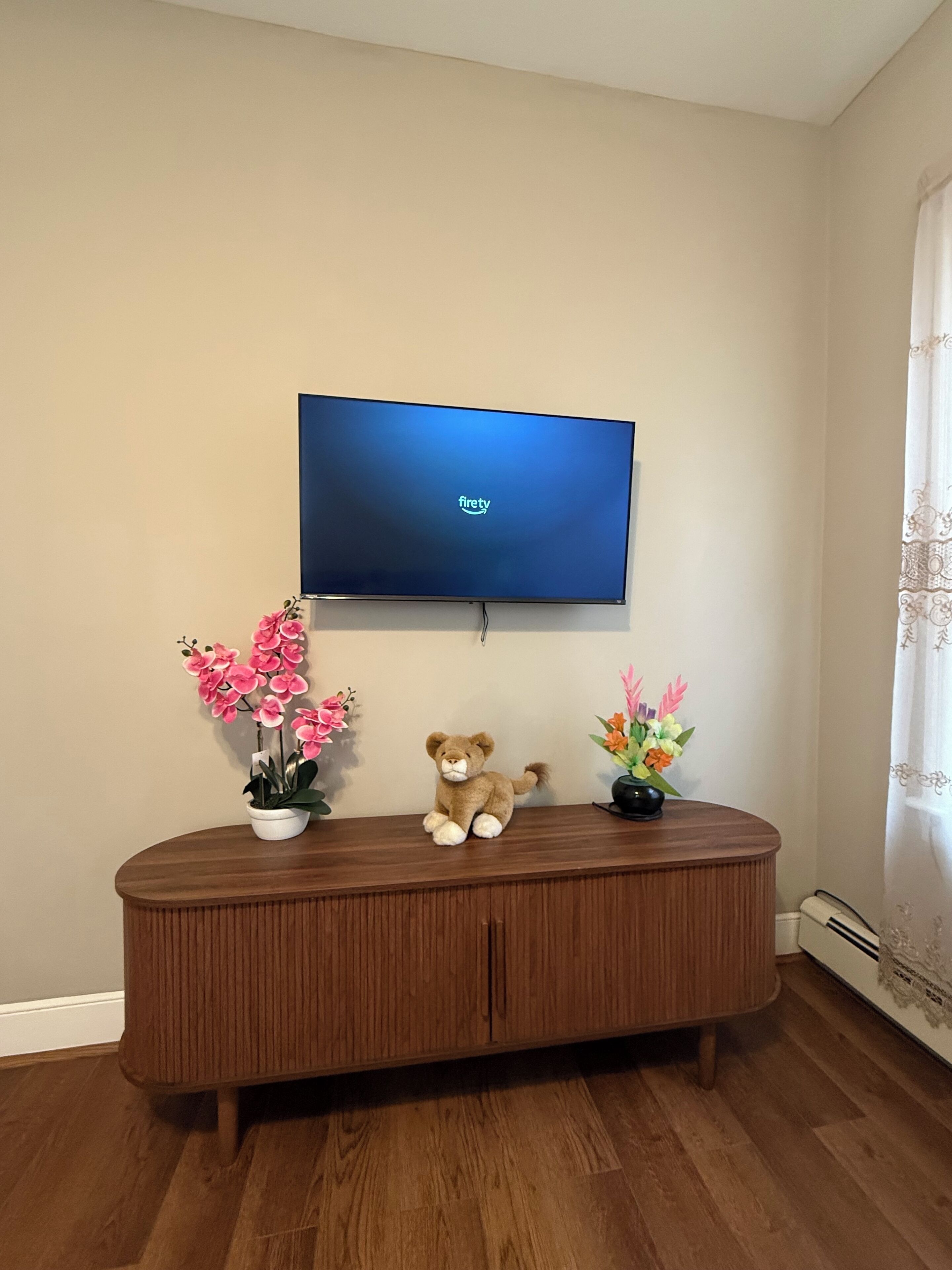 A television mounted on a wall, a wooden console table with a teddy bear and potted plants, and a wooden floor.